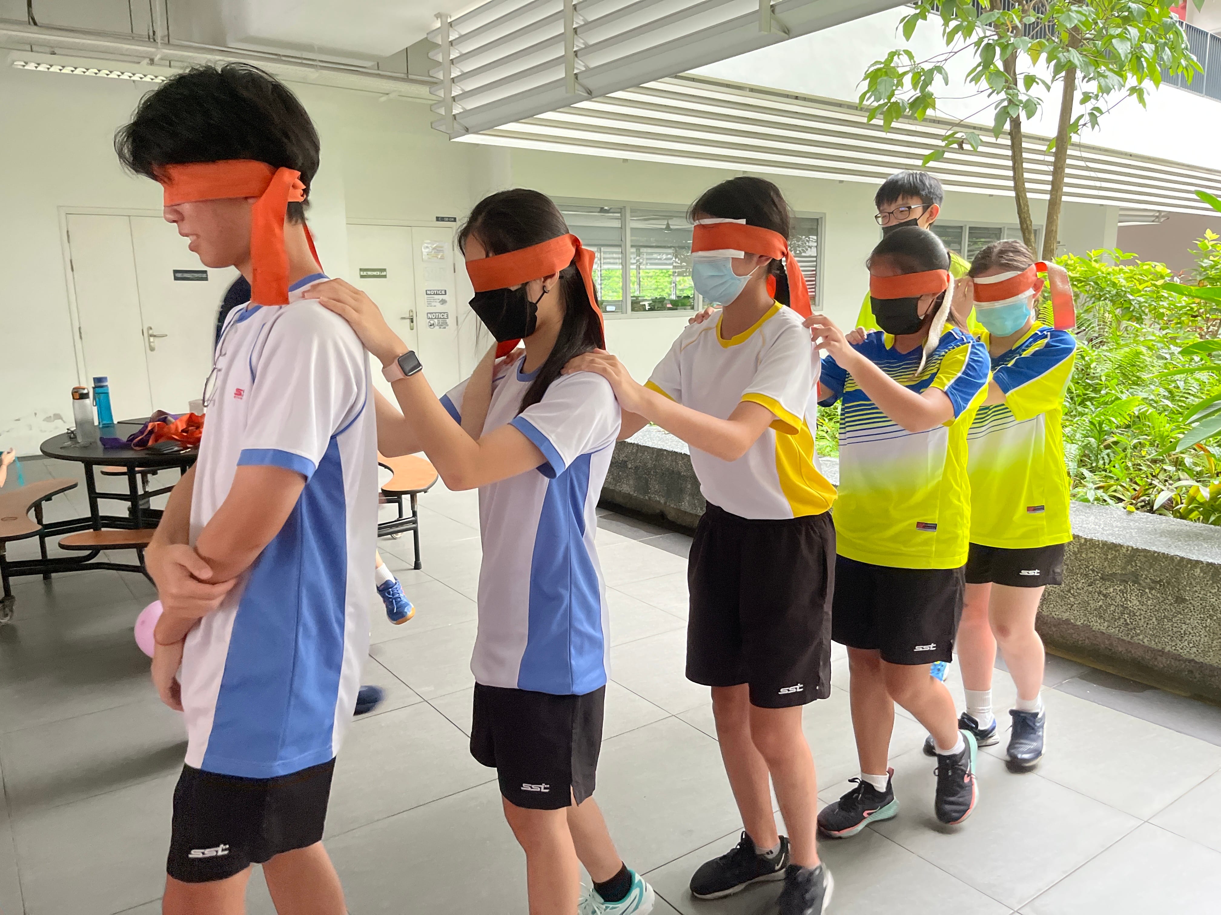 Students in sports uniforms form a chain, most wearing orange blindfolds, each placing hands on the shoulders ahead.